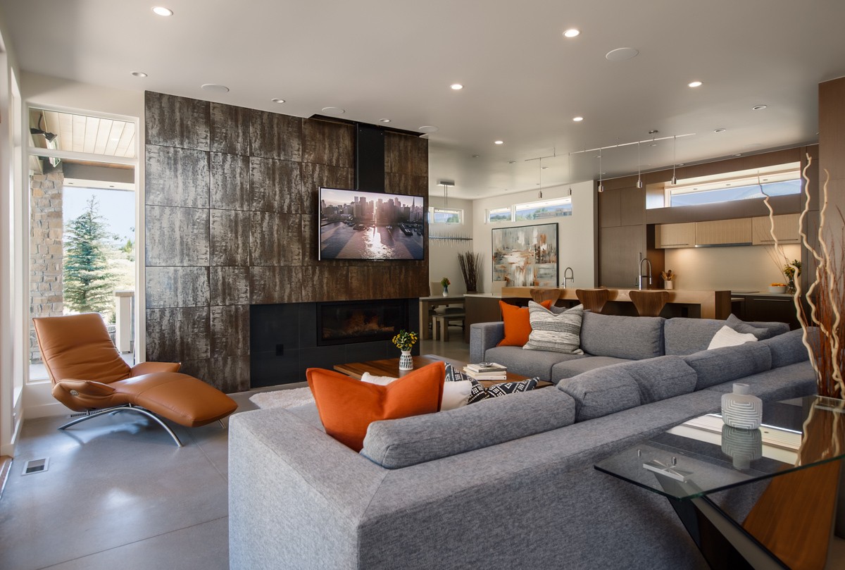 A modern living room with a wall-mounted TV, comfortable seating including a gray sectional sofa and a tan recliner, a cozy fireplace, and an open concept leading to the kitchen area. 