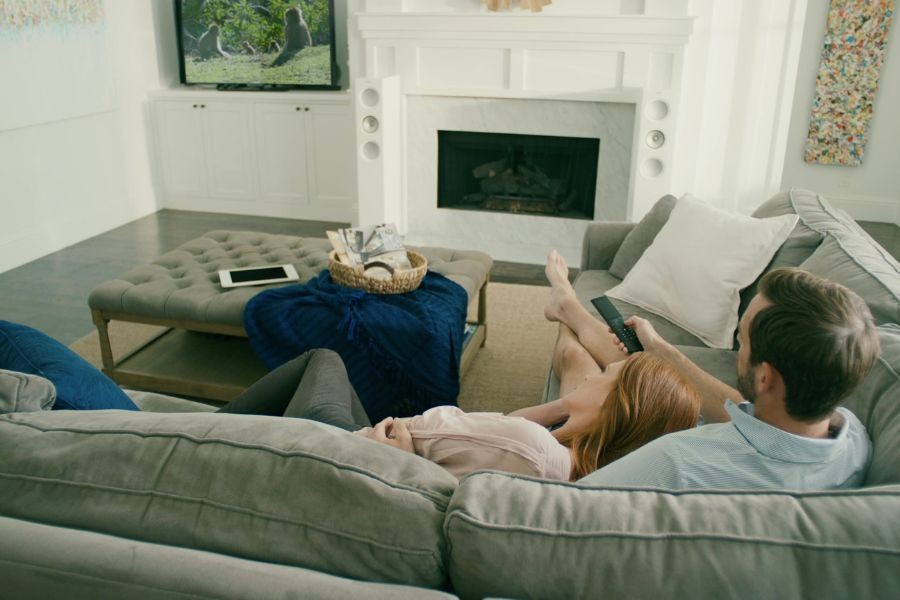 Couple sitting on the couch using a Savant remote, with a Savant tablet on the ottoman, watching television. 