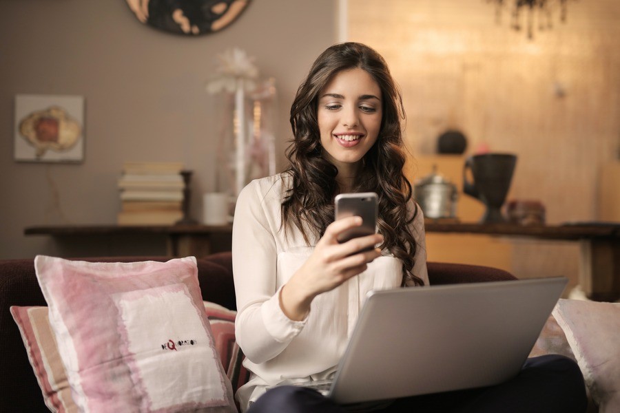 Woman sitting on her couch with her phone, a laptop open on her lap.